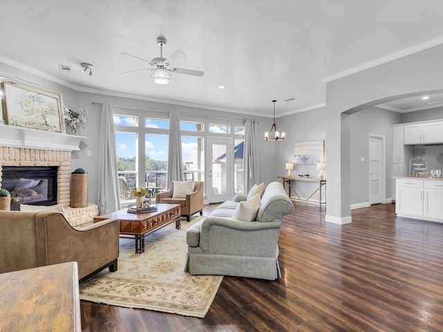 living room with a brick fireplace, dark wood-type flooring, ceiling fan with notable chandelier, and ornamental molding