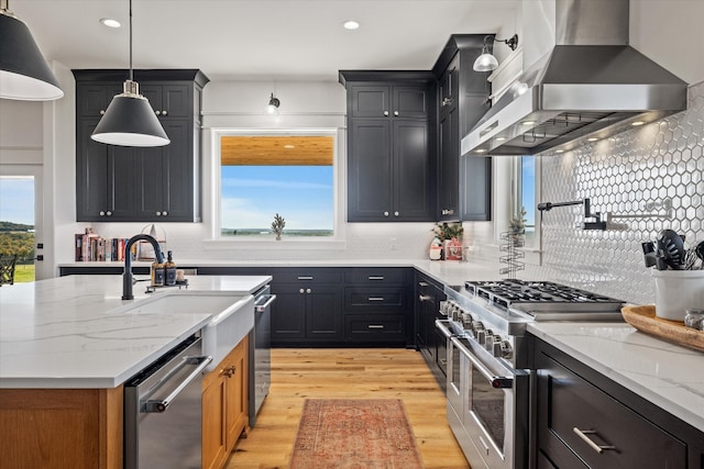 kitchen featuring stainless steel appliances, wall chimney range hood, light stone counters, pendant lighting, and light hardwood / wood-style floors