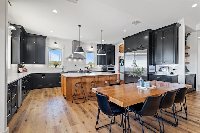 kitchen with pendant lighting, a center island, wall chimney range hood, light hardwood / wood-style floors, and stainless steel built in refrigerator