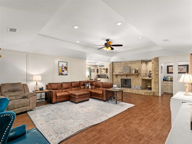 living room with dark hardwood / wood-style flooring, a tray ceiling, a brick fireplace, and ceiling fan