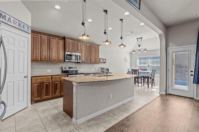 kitchen featuring light wood-type flooring, light stone counters, stainless steel appliances, decorative light fixtures, and a center island with sink