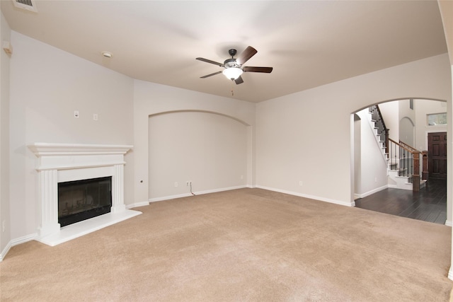 unfurnished living room featuring stairway, carpet floors, arched walkways, a glass covered fireplace, and a ceiling fan