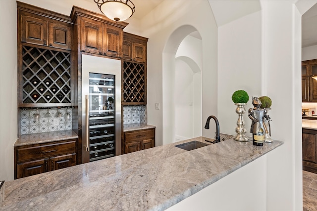 bar with sink, tasteful backsplash, light stone countertops, beverage cooler, and dark brown cabinetry