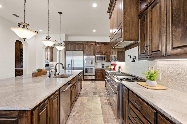 kitchen with built in appliances, tasteful backsplash, sink, decorative light fixtures, and a large island with sink