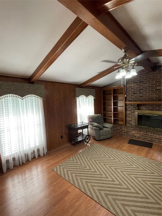 unfurnished living room with beam ceiling, light wood-style flooring, a brick fireplace, wood walls, and ceiling fan