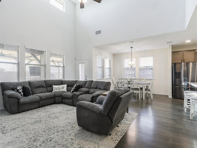 living room featuring dark wood-type flooring, a towering ceiling, a healthy amount of sunlight, and ceiling fan with notable chandelier