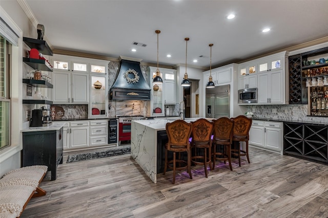 kitchen with premium range hood, a center island with sink, white cabinets, hanging light fixtures, and built in appliances