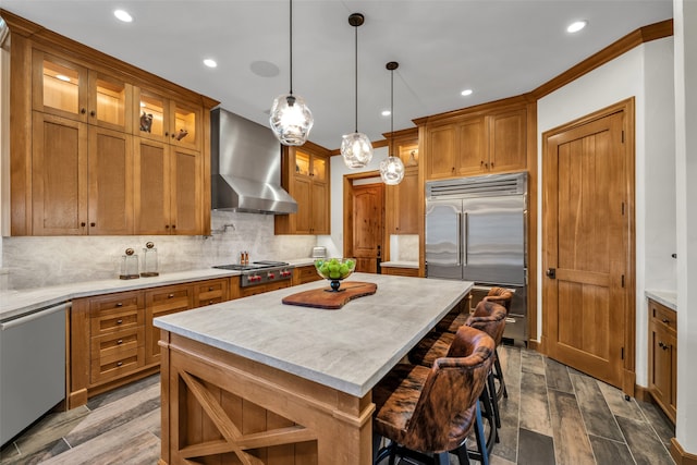 kitchen with a center island, wall chimney range hood, hanging light fixtures, appliances with stainless steel finishes, and wood-type flooring