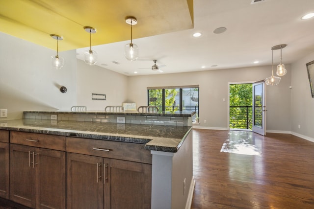 kitchen with dark wood-type flooring, dark stone counters, hanging light fixtures, ceiling fan, and a breakfast bar area