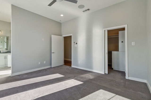 unfurnished bedroom featuring a walk in closet, ensuite bathroom, dark carpet, and ceiling fan