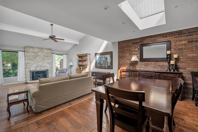 dining room featuring a stone fireplace, vaulted ceiling with skylight, ceiling fan, wood-type flooring, and brick wall