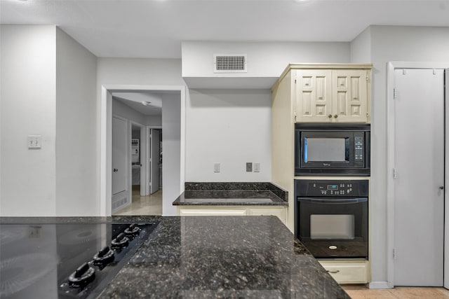 kitchen featuring black appliances, cream cabinetry, light tile patterned floors, and dark stone counters