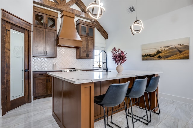 kitchen featuring decorative backsplash, premium range hood, light stone counters, a kitchen island with sink, and pendant lighting