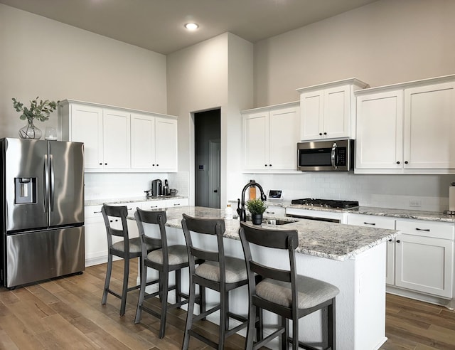 kitchen with sink, appliances with stainless steel finishes, light stone countertops, a kitchen island with sink, and white cabinets