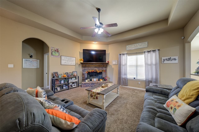 carpeted living room featuring a tray ceiling, a brick fireplace, and ceiling fan