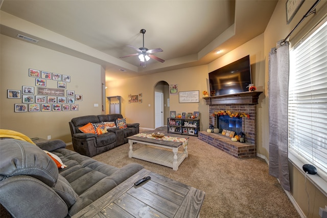 carpeted living room with ceiling fan, a tray ceiling, and a brick fireplace