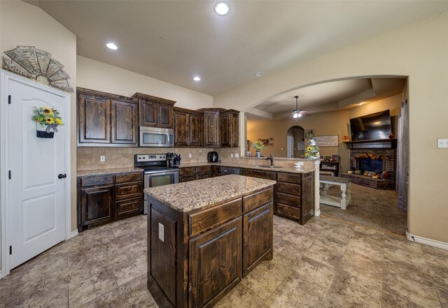 kitchen featuring kitchen peninsula, a brick fireplace, dark brown cabinetry, stainless steel appliances, and a center island