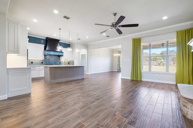 kitchen with white cabinetry, hanging light fixtures, a kitchen island with sink, custom range hood, and ornamental molding