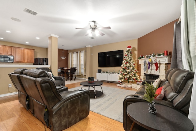 living room featuring light wood-type flooring, a stone fireplace, ceiling fan, and ornate columns