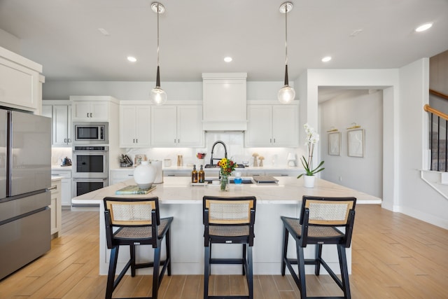 kitchen featuring appliances with stainless steel finishes, a kitchen island with sink, pendant lighting, white cabinets, and light hardwood / wood-style floors