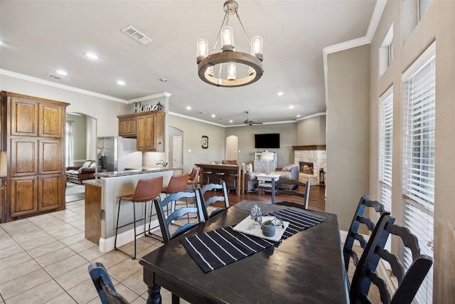 dining area with ceiling fan with notable chandelier, a fireplace, light tile patterned flooring, and crown molding