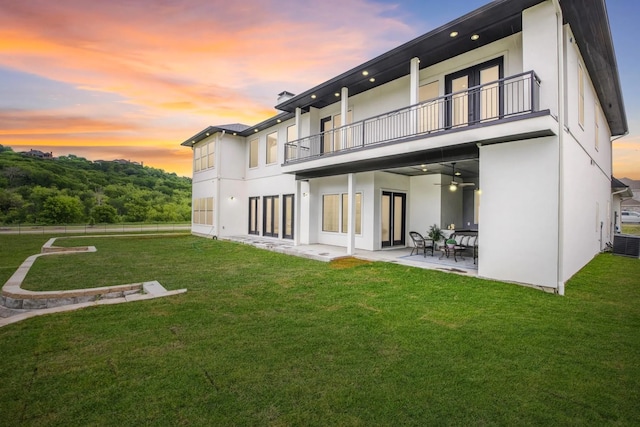 back house at dusk with a lawn, ceiling fan, a balcony, central AC unit, and a patio