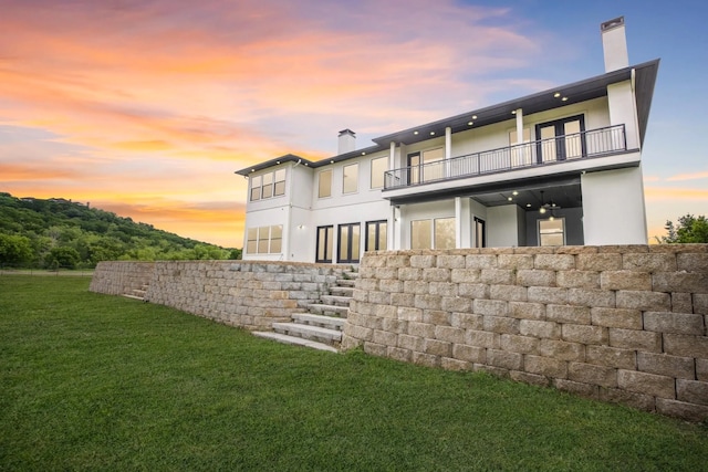 back house at dusk featuring a lawn and a balcony