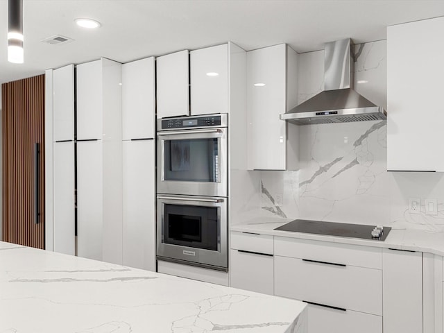 kitchen with light stone counters, stainless steel double oven, black electric cooktop, wall chimney range hood, and white cabinets