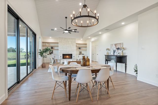 dining area featuring built in shelves, ceiling fan with notable chandelier, light hardwood / wood-style flooring, high vaulted ceiling, and a stone fireplace