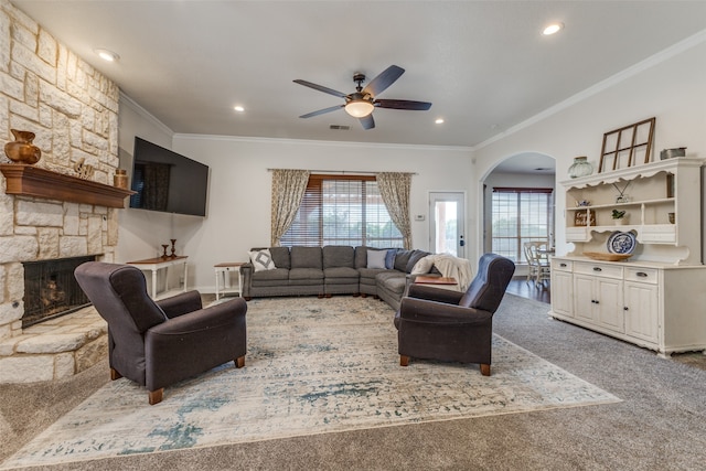 carpeted living room featuring a stone fireplace, ceiling fan, and crown molding