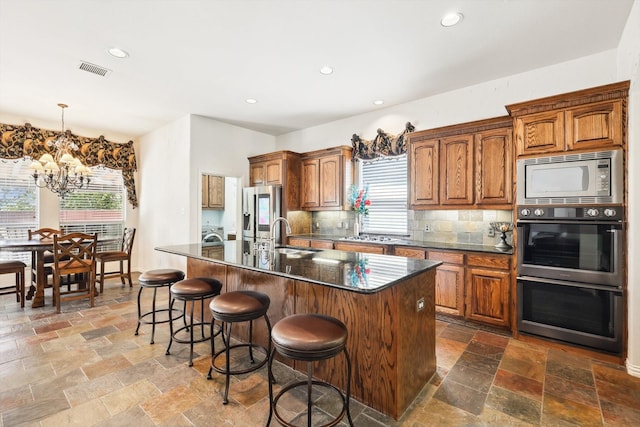 kitchen featuring sink, stainless steel appliances, a notable chandelier, backsplash, and an island with sink