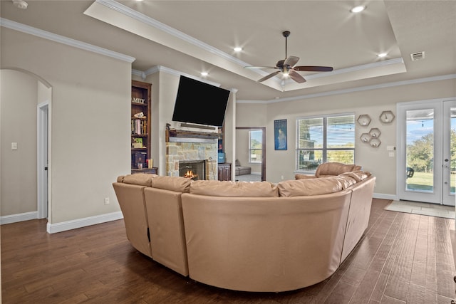 living room with ceiling fan, a raised ceiling, a stone fireplace, dark hardwood / wood-style floors, and ornamental molding