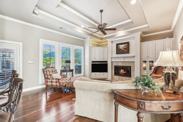 living room with a tile fireplace, ceiling fan, dark wood-type flooring, crown molding, and a tray ceiling