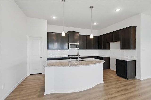 kitchen with stainless steel microwave, a sink, light wood-type flooring, light stone counters, and a kitchen island with sink