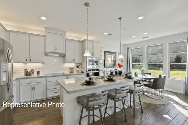 kitchen with appliances with stainless steel finishes, dark hardwood / wood-style floors, hanging light fixtures, and a kitchen island with sink