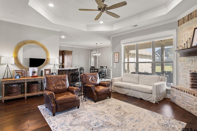 living room with a raised ceiling, a stone fireplace, dark hardwood / wood-style floors, crown molding, and ceiling fan with notable chandelier