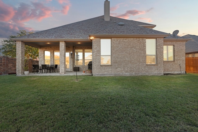 back of property at dusk featuring a lawn, roof with shingles, fence, a patio area, and brick siding