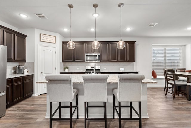 kitchen with an island with sink, decorative light fixtures, light stone countertops, appliances with stainless steel finishes, and a kitchen bar