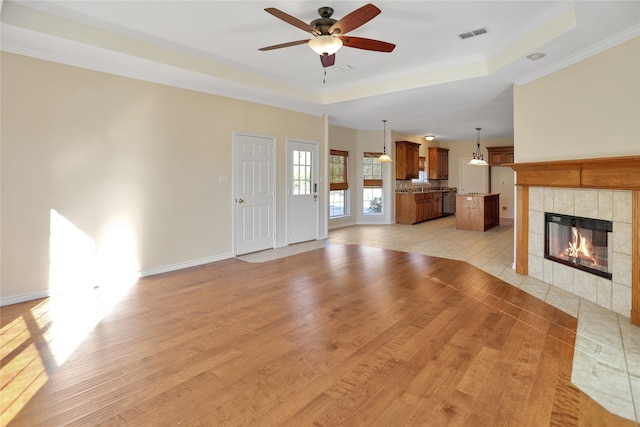 unfurnished living room with a tiled fireplace, ornamental molding, ceiling fan, a raised ceiling, and light wood-type flooring