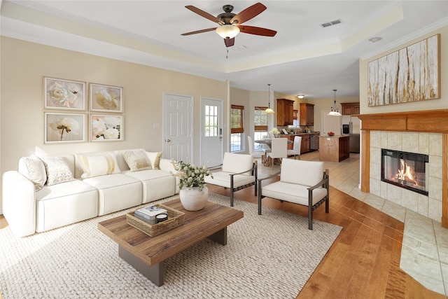 living room featuring crown molding, a raised ceiling, ceiling fan, a tiled fireplace, and hardwood / wood-style floors