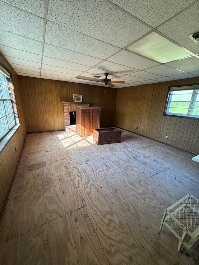 unfurnished living room featuring a fireplace, ceiling fan, a drop ceiling, and wood walls