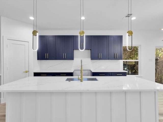 kitchen featuring hanging light fixtures, light stone counters, blue cabinets, a center island with sink, and light wood-type flooring