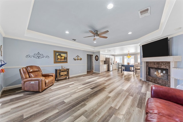 living room with a high end fireplace, light hardwood / wood-style flooring, ceiling fan, ornamental molding, and a tray ceiling