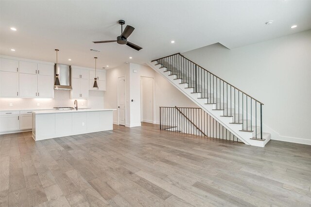 kitchen featuring a kitchen island with sink, wall chimney range hood, ceiling fan, light hardwood / wood-style floors, and white cabinetry