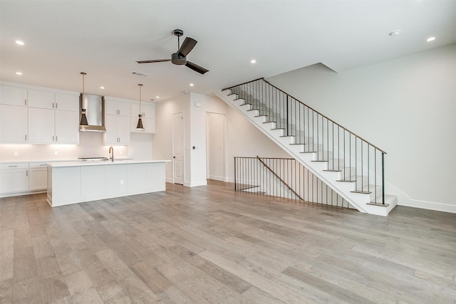 kitchen featuring a kitchen island with sink, wall chimney range hood, ceiling fan, light hardwood / wood-style floors, and white cabinetry