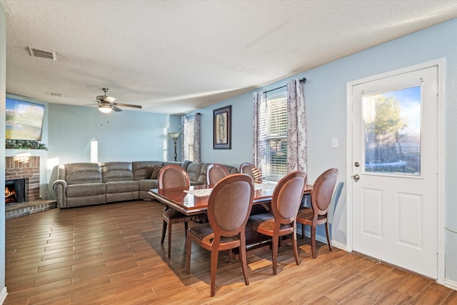 dining area featuring ceiling fan, a textured ceiling, a brick fireplace, and light wood-type flooring