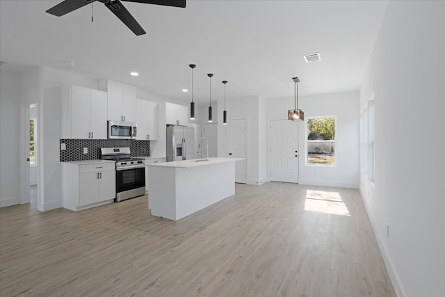 kitchen with stainless steel appliances, pendant lighting, light hardwood / wood-style flooring, white cabinets, and an island with sink