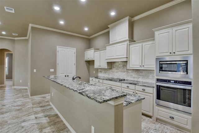 kitchen featuring light stone countertops, appliances with stainless steel finishes, tasteful backsplash, a center island with sink, and white cabinetry