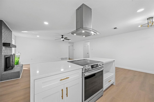 kitchen with stainless steel range with electric stovetop, light hardwood / wood-style flooring, a brick fireplace, island exhaust hood, and white cabinets