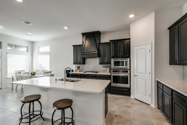 kitchen with sink, a breakfast bar area, a kitchen island with sink, custom range hood, and appliances with stainless steel finishes